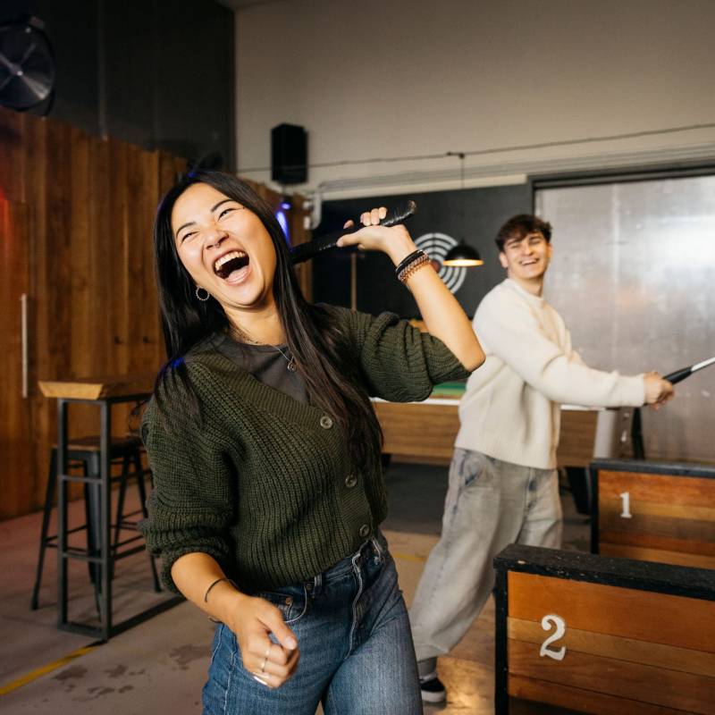 Happy young woman throwing an axe in a axe throwing room. Group of friends enjoying at a Axe throwing room.