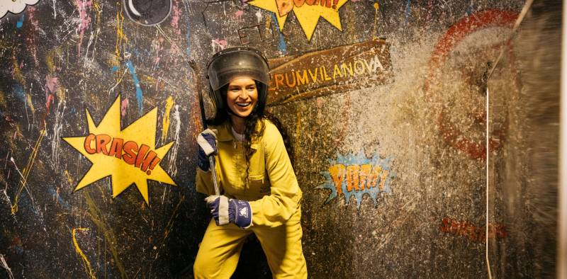 Young woman wearing safety clothes and helmet breaking things with a metal bar in a rage room. Young woman destroying a TV in a fury room